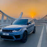 A sleek blue Range Rover parked on a curvy bridge at sunset, with city skyscrapers in the background and a warm orange sky.
