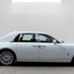 Side view of a sleek, luxury white Rolls-Royce with chrome accents parked in a minimalist studio setting.